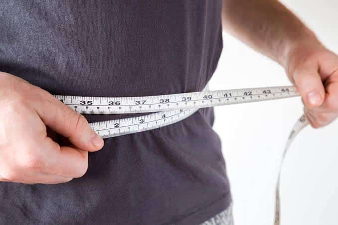 Overweight man measuring his waist circumference with a tailor's measuring tape to assess abdominal obesity.