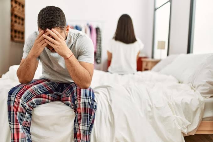 Distressed Indian man experiencing erectile dysfunction sitting on bed near supportive partner