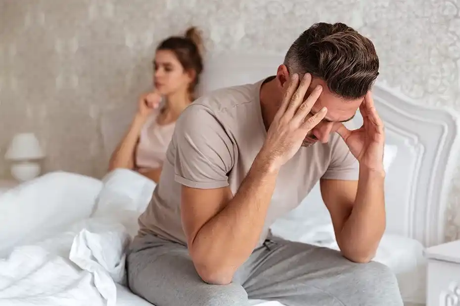 Indian man with erectile dysfunction feeling distressed while partner sits on bed, illustrating relationship challenges caused by erectile dysfunction.